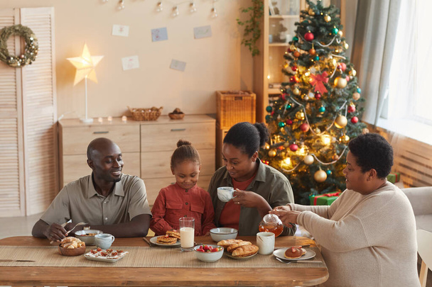 Retrato cálido tonificado de la feliz familia afroamericana disfrutando de té y aperitivos mientras celebra la Navidad en casa en el acogedor interior del hogar, espacio para copiar - Foto, imagen
