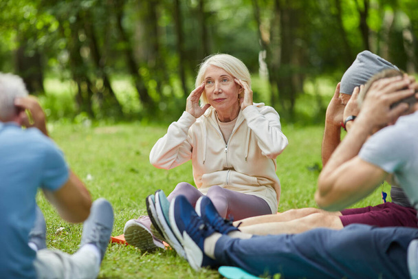 Група сучасних людей похилого віку зібралася в парку, сидячи на килимках на траві, роблячи шию розслаблюючою вправою
 - Фото, зображення