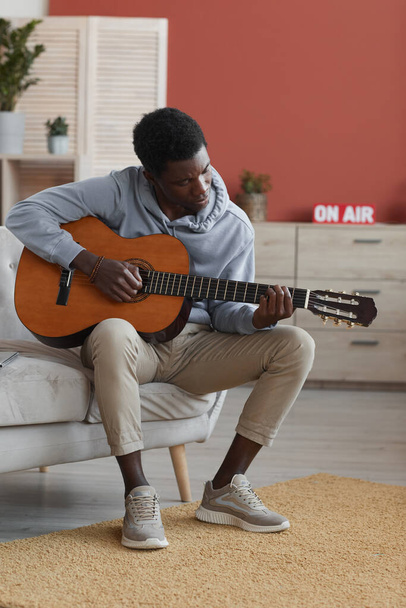 Vertical full length portrait of young African-American man playing acoustic guitar while sitting on couch at home - Φωτογραφία, εικόνα