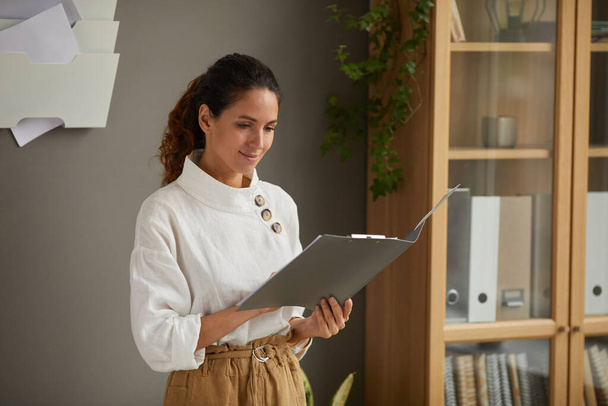 Waist up portrait of smiling successful businesswoman reading documents in open fol der while standing by grey wall in office, copy space - Φωτογραφία, εικόνα
