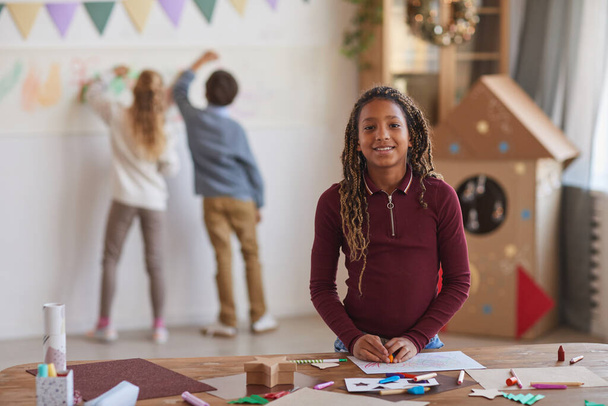 Талия портрет улыбающейся афро-американской девушки, смотрящей в камеру, стоя у стола и наслаждаясь классом рисования в школе, копировать пространство - Фото, изображение