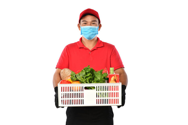 赤い制服を着た幸せな若いアジアの配達人、医療面マスクは白い背景に隔離された手の中に食料品箱を運ぶ - 写真・画像