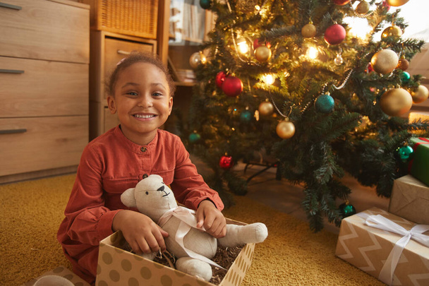 Portret szczęśliwej Afroamerykanki otwierającej prezenty świąteczne siedząc przy drzewie w domu i uśmiechając się do aparatu fotograficznego, przestrzeń kopiująca - Zdjęcie, obraz