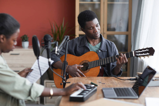 Ev kayıt stüdyosunda müzik bestelerken akustik gitar çalan genç Afro-Amerikan bir adamın portresi. - Fotoğraf, Görsel