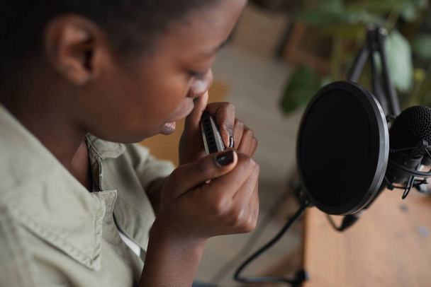 Высокоугольный крупный план молодой афроамериканки, играющей на губной гармошке под микрофон и компилирующей музыку в домашней студии звукозаписи, копировальное пространство - Фото, изображение