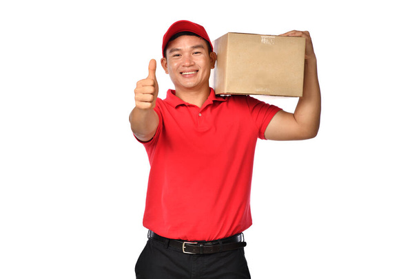 Ασιάτης διανομέας με κόκκινη στολή με χάρτινο κουτί δεμάτων και δείχνοντας τον αντίχειρά του επάνω απομονωμένος σε λευκό φόντο                                                                                                                          - Φωτογραφία, εικόνα