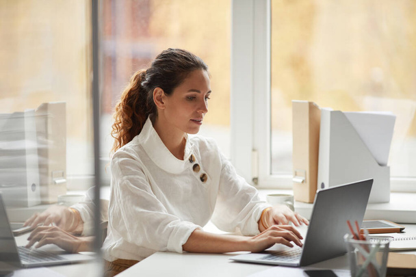 Portret van een succesvolle zakenvrouw met behulp van laptop tijdens het zitten aan het bureau door het raam en genieten van het werk op kantoor, kopieer ruimte - Foto, afbeelding