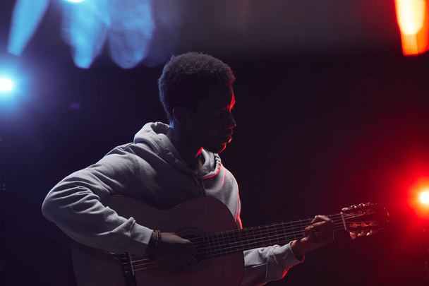 Side view outline of young African-American man playing acoustic guitar on stage in colored lights, copy space - Photo, Image