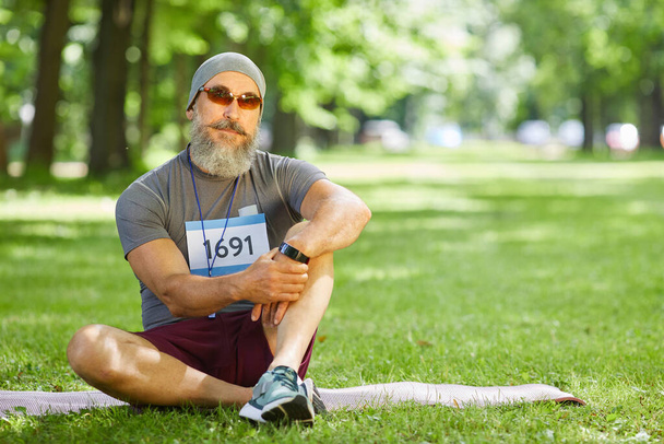 Portrét stylového staršího muže s vousy na obličeji, který se účastní letního maratonu, odpočatý na trávě - Fotografie, Obrázek
