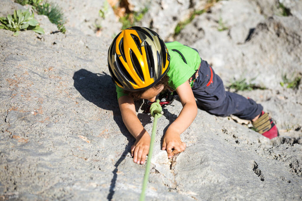 The child is climbing on a natural terrain. A boy in helmet climbs a rock on a background of mountains. Extreme hobby. Athletic kid trains to be strong. Rock climbing safety. - Photo, Image