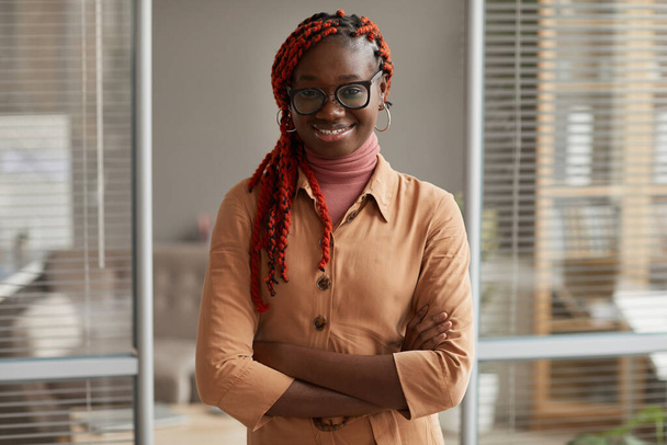 Portrait taille haute de la jeune femme d'affaires afro-américaine souriant à la caméra tout en posant avec confiance debout les bras croisés dans le bureau, espace de copie - Photo, image
