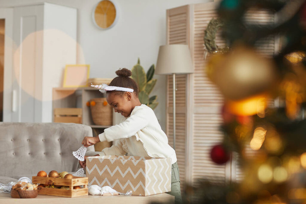 Вид сбоку портрет милой афроамериканской девушки, открывающий подарочную коробку в уютном интерьере дома, рождественский сюрприз, копировальное пространство - Фото, изображение