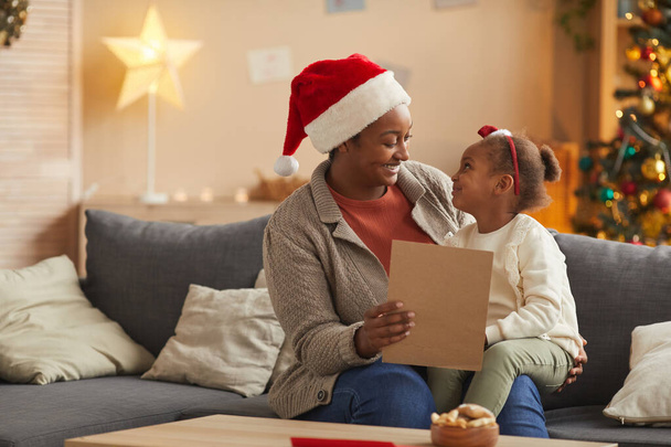 自宅でクリスマスシーズンを楽しみながらかわいい女の子とサンタにアフリカ系アメリカ人の母親読書手紙の肖像画、コピースペース - 写真・画像