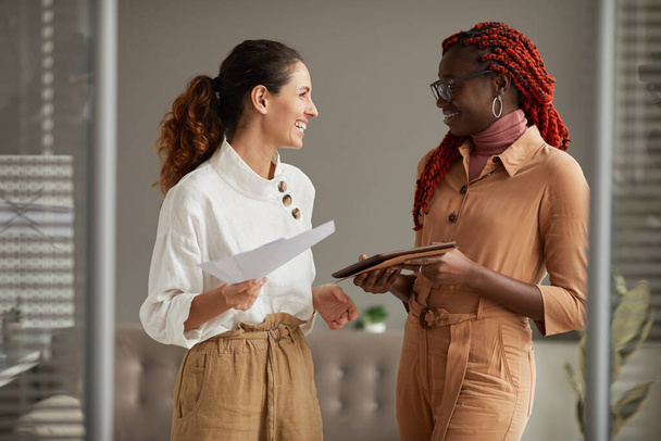Waist up portrait of two successful young businesswomen smiling cheerfully looking at each other while standing in modern office interior, copy space - Photo, Image