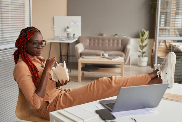 若いアフリカ系アメリカ人女性の完全な長さの肖像画テイクアウト食品を食べ、自宅のオフィスでリラックスしながら、ノートパソコンの画面を見て、スペースをコピー - 写真・画像