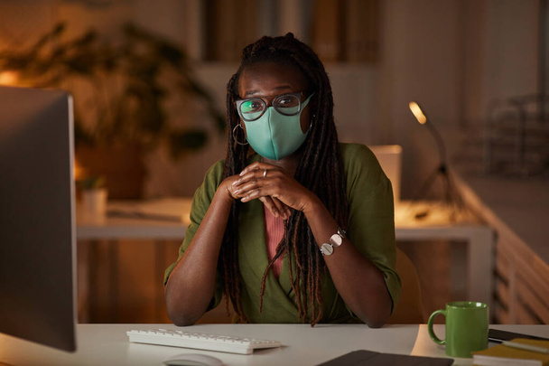 Portret van een jonge Afro-Amerikaanse vrouw met masker en bril die 's nachts naar de camera kijkt terwijl ze met een pc op kantoor werkt, kopieer ruimte - Foto, afbeelding