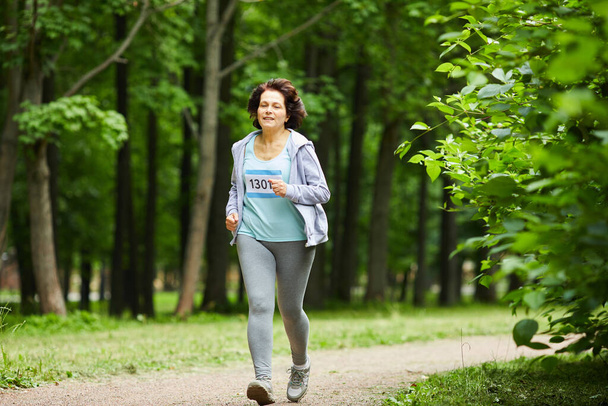 Full body shot of modern mature woman with brown hair running marathon race in forest park - Foto, imagen