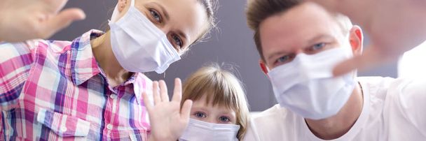 Man, woman and little girl in protective medical masks on face waving in front of laptop camera - 写真・画像