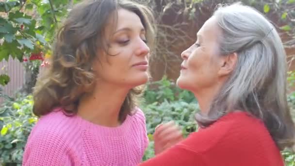 Senior μητέρα με γκρίζα μαλλιά με την ενήλικη κόρη της κοιτάζοντας την κάμερα στον κήπο και αγκαλιάζοντας ο ένας τον άλλον κατά τη διάρκεια ηλιόλουστη μέρα σε εξωτερικούς χώρους - Πλάνα, βίντεο