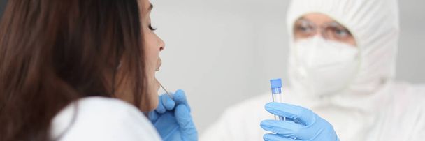 Dokter in witte anti-pest pak neemt uitstrijkje van zijn keel met wattenstaafje in reageerbuis in appartement - Foto, afbeelding