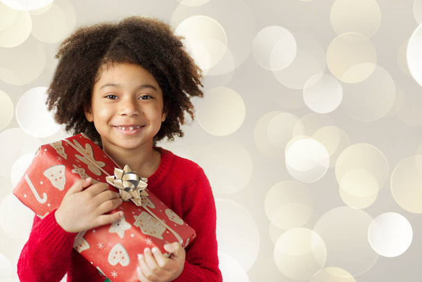 Gülümseyen, kıvırcık saçlı küçük bir kızın portresi. Elinde bir Noel hediyesi var.  - Fotoğraf, Görsel