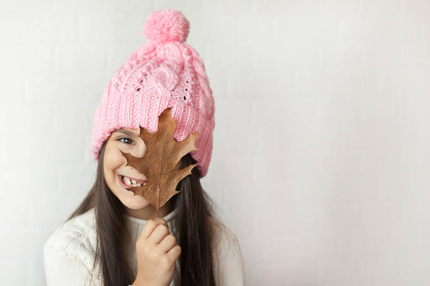 retrato de una hermosa niña en un sombrero de punto rosa y con hoja de otoño en las manos - Foto, Imagen