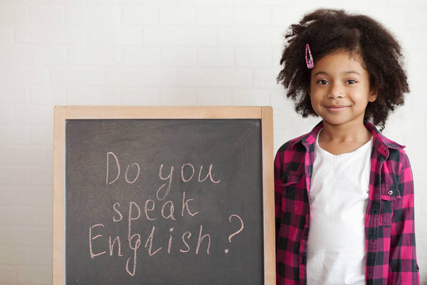 Счастливая школьница держит в руках доску с текстом: "Вы говорите по-английски?? - Фото, изображение