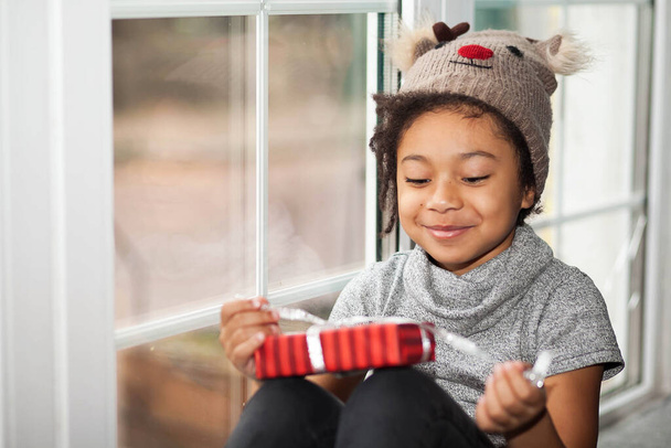 πορτρέτο ενός χαμογελαστού κοριτσιού με καπέλο με το φίμωτρο του ελαφιού να ανοίγει ένα χριστουγεννιάτικο δώρο δίπλα στο παράθυρο - Φωτογραφία, εικόνα