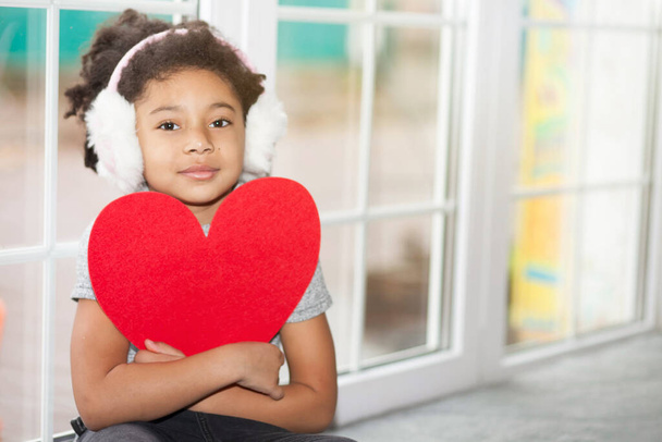 Bambina afroamericana con cuffie invernali vicino alla finestra con il cuore rosso - Foto, immagini