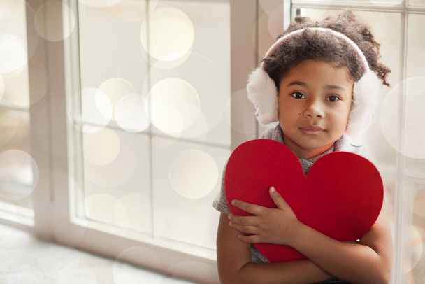 African American κοριτσάκι με ακουστικά χειμώνα από το παράθυρο κρατώντας κόκκινη καρδιά - Φωτογραφία, εικόνα