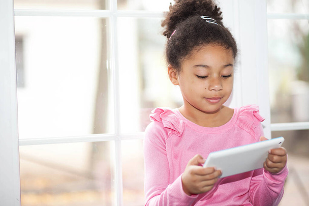 carino bambina utilizzando un tablet mentre seduto vicino alla finestra a casa - Foto, immagini