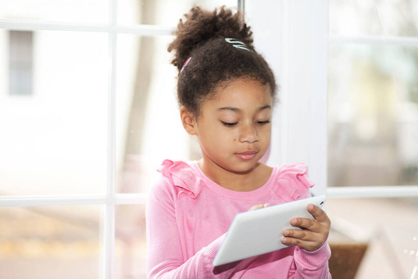 aranyos kislány használ egy tablettát, miközben otthon ül az ablaknál - Fotó, kép