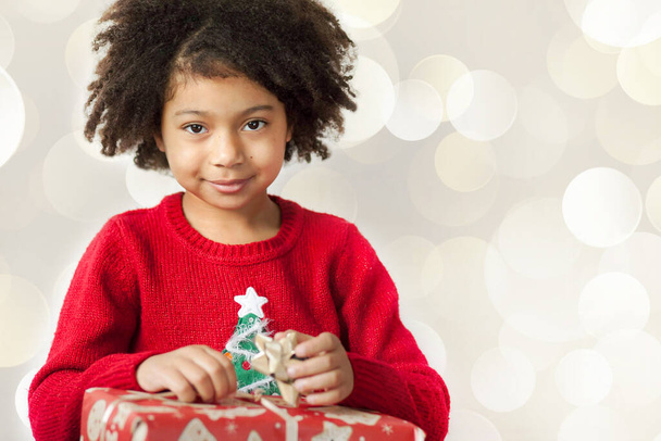 Porträt eines lächelnden kleinen Mädchens mit lockigem Haar, das ein Weihnachtsgeschenk in der Hand hält  - Foto, Bild