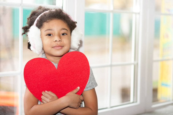 Niña afroamericana con auriculares de invierno junto a la ventana sosteniendo el corazón rojo - Foto, imagen