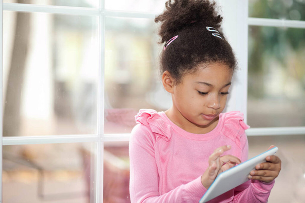 carino bambina utilizzando un tablet mentre seduto vicino alla finestra a casa - Foto, immagini