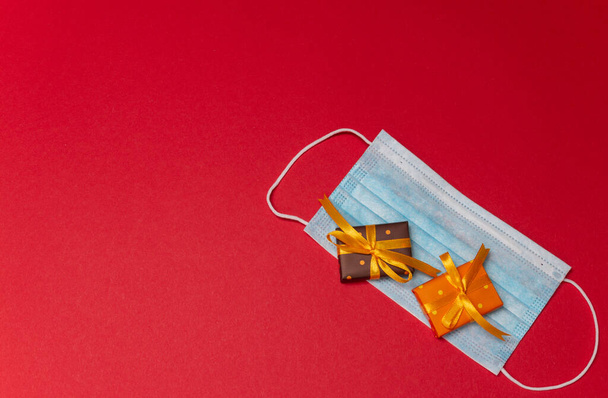 Modrá lékařská maska na vrchu se dvěma krabičkami v barevném papíru s puntíky svázané žlutou stuhou s dárkem na červeném pozadí. Dárek nebo překvapení k Vánocům, narozeninám nebo k výročí.  - Fotografie, Obrázek