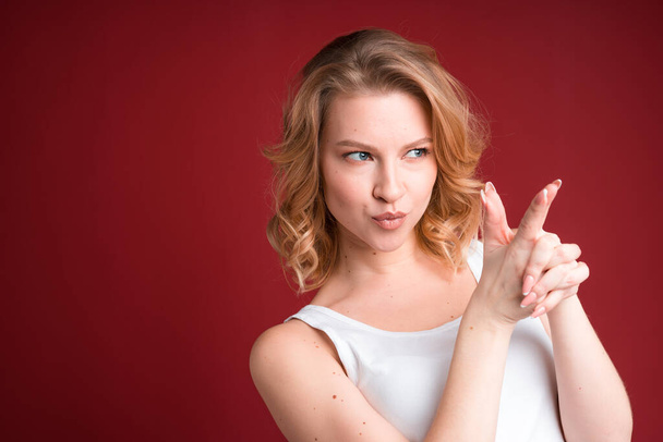 Blond woman with curly hair showing finger gun in white tank top on red background. - Foto, imagen