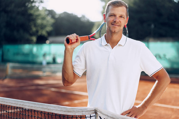 Portrait of positive male tennis player with racket standing at clay court - Foto, afbeelding