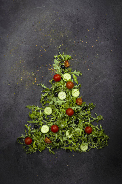 Rugula di verdure fresche, pomodoro, cetriolo, verdure, grani di pepe e olive su uno sfondo strutturato grigio scuro sotto forma di albero di Natale. Vista dall'alto. - Foto, immagini