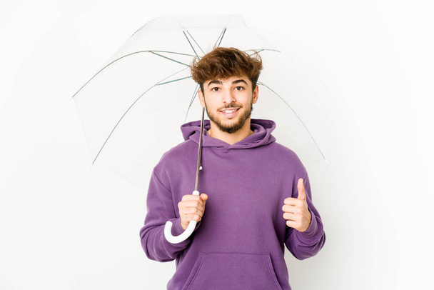 Young arab man holding an umbrella smiling and raising thumb up - Photo, Image