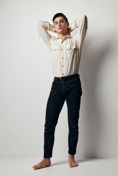 A handsome guy in a white shirt and trousers holds his hands behind his head on a light background - Foto, Imagem
