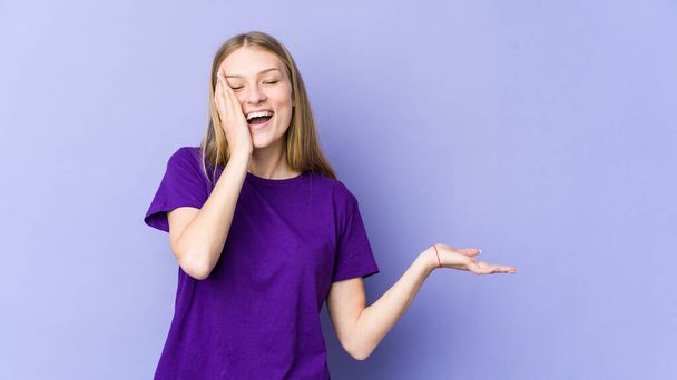 Young blonde woman isolated on purple background holds copy space on a palm, keep hand over cheek. Amazed and delighted. - Photo, Image