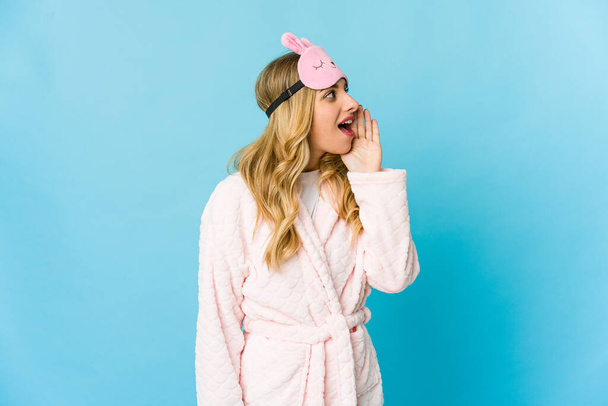 Young blonde caucasian woman wearing a pajama shouting and holding palm near opened mouth. - Foto, Bild