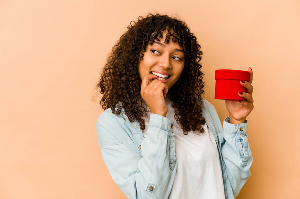 Junge afrikanisch-amerikanische Afro-Frau hält ein Valentinstagsgeschenk in der Hand und denkt entspannt über etwas nach, das einen Kopierraum betrachtet. - Foto, Bild