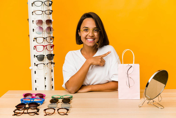 Junge hispanische Frau probiert eine Brille an, lächelt isoliert und zeigt zur Seite, zeigt etwas im leeren Raum. - Foto, Bild