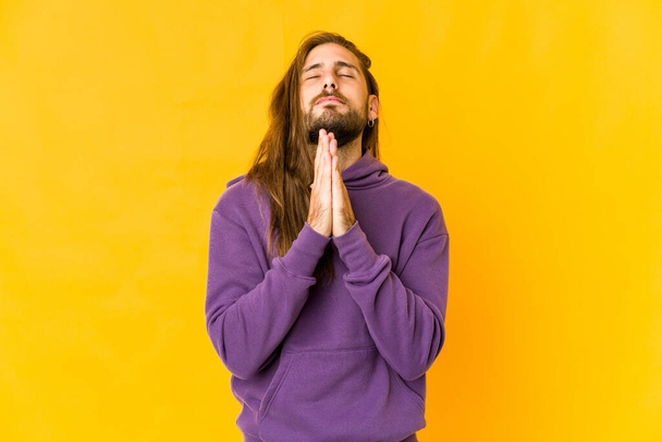 Молодий чоловік з довгим волоссям дивиться, тримаючи руки в молитві біля рота, відчуває себе впевнено
. - Фото, зображення
