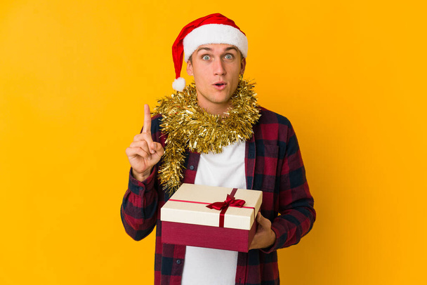 Mladý běloch s vánočním kloboukem drží dárek izolovaný na žlutém pozadí s nějakou skvělou myšlenkou, koncept kreativity. - Fotografie, Obrázek