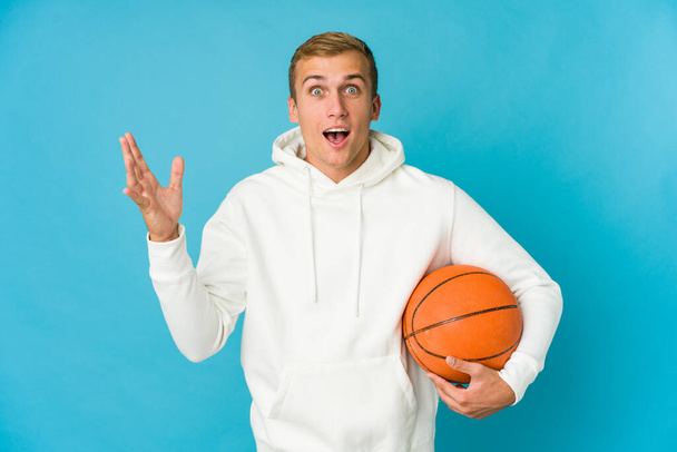 Nuori valkoihoinen mies pelaa koripalloa eristetty sinisellä taustalla saavat miellyttävä yllätys, innoissaan ja nostamalla kädet. - Valokuva, kuva