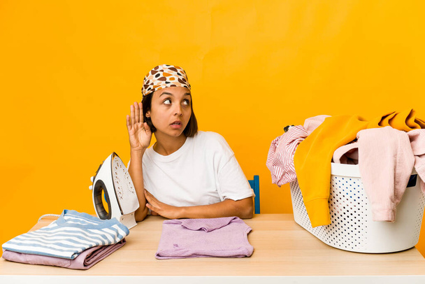 Mujer hispana joven planchando ropa aislada tratando de escuchar un chisme. - Foto, imagen