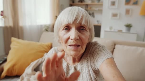 Oudere grijsharige vrouw zwaaiend naar de camera en sprekend op online videogesprek terwijl ze op de bank zit in de woonkamer - Video